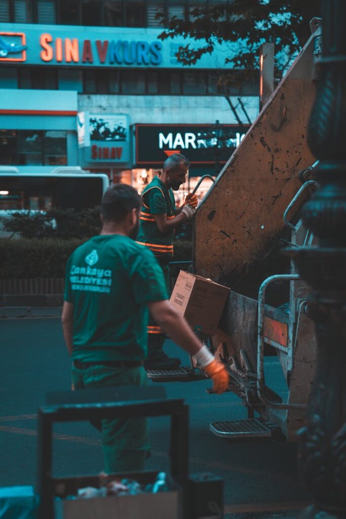 Two workers operate a garbage truck in a city street at night, wearing uniforms.